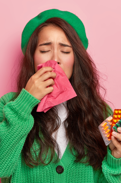 Unhappy brunette girl suffers from flu symptoms, rubs nose with handkerchief, has cold, holds pills, wears green jumper and cap, isolated on pink wall. Seasoanl disease, running nose and medicaments